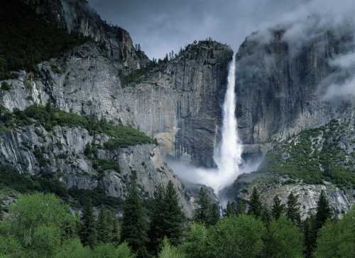 A majestic waterfall cascades down rocky cliffs, surrounded by lush greenery and misty clouds in a dramatic landscape.