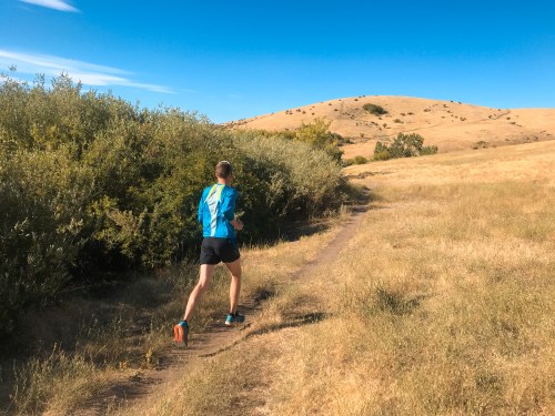 A person runs along a dirt path through grassy fields and shrubs, with a hill in the background under a clear blue sky.
