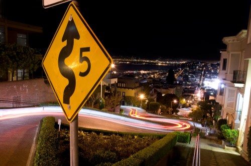 A winding road sign at night, with city lights and a bay visible in the background.