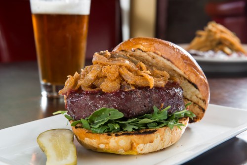 A juicy burger topped with crispy onions and greens, served with a pickle and a glass of beer in the background.