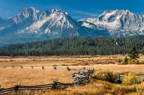 A scenic view of mountains with snow-capped peaks, surrounded by a grassy meadow and wooden fences under a clear blue sky.