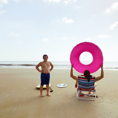A boy stands on the beach while a person in a chair holds a large pink float, with surfboards nearby and ocean in the background.