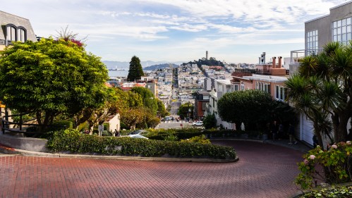 A winding street lined with greenery, leading down to a cityscape with hills and a distant tower under a blue sky.