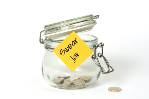 A glass jar labeled "Swear jar" with coins inside and a lid slightly open, sitting on a white background.
