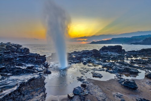 A natural geyser erupts from rocky shorelines at sunset, with waves crashing against the rocks and a colorful sky.