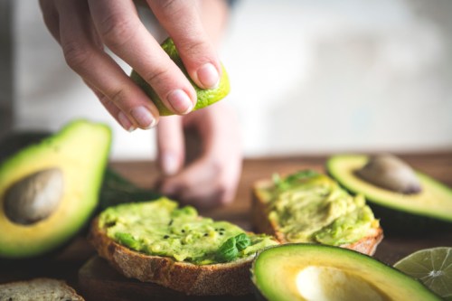 A hand squeezing lime over avocado toast, surrounded by fresh avocados on a wooden surface.