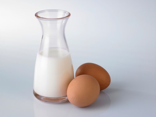 A glass pitcher of milk next to two brown eggs on a light surface.