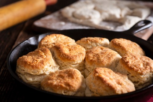 Freshly baked biscuits in a cast iron skillet, with a rolling pin and dough in the background.