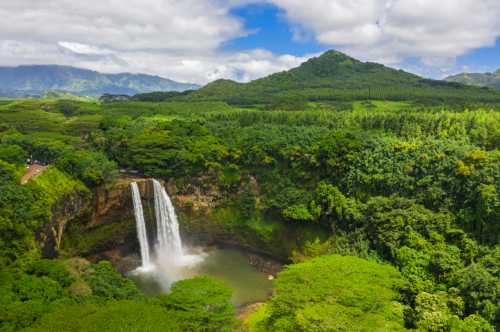 A stunning waterfall cascades into a lush green valley surrounded by mountains and dense foliage under a cloudy sky.