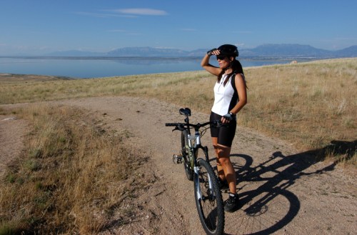 A woman in cycling gear stands beside her bike, looking out over a scenic landscape with mountains and a lake.
