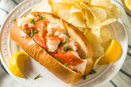 A lobster roll filled with lobster meat, served with potato chips and lemon wedges on a white plate.