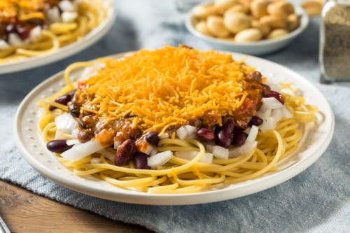 A plate of spaghetti topped with chili, kidney beans, diced onions, and shredded cheddar cheese, with snacks in the background.