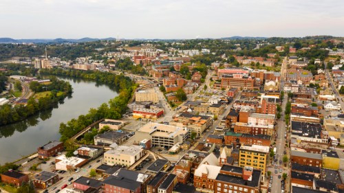 Aerial view of a riverside town with buildings, roads, and green hills in the background under a cloudy sky.