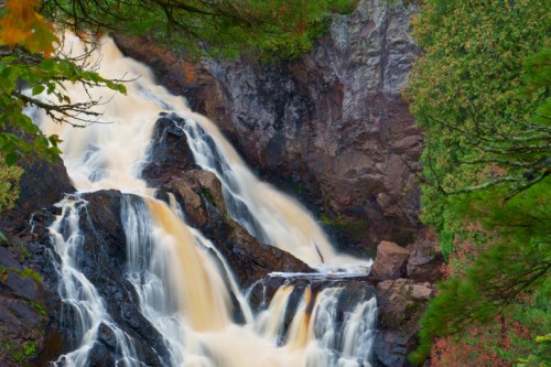 A cascading waterfall flows over rocky cliffs, surrounded by lush green trees and vibrant autumn foliage.