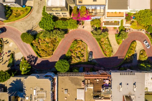 Aerial view of a winding road surrounded by greenery and colorful flowers, with buildings and parked cars nearby.