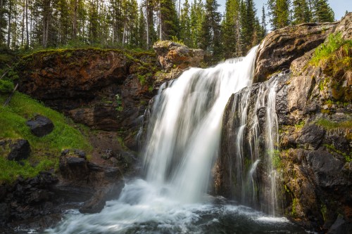 A serene waterfall cascading over rocks, surrounded by lush greenery and tall trees in a tranquil natural setting.