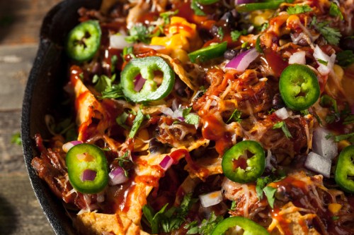 A close-up of a sizzling plate of nachos topped with cheese, jalapeños, onions, and barbecue sauce.