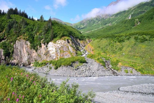 A serene landscape featuring a winding river surrounded by lush green hills and rocky cliffs under a clear blue sky.