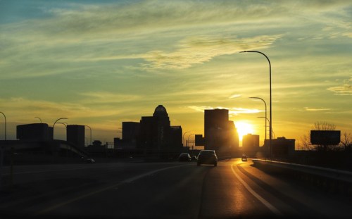 Silhouette of a city skyline at sunset, with a winding road and glowing streetlights in the foreground.