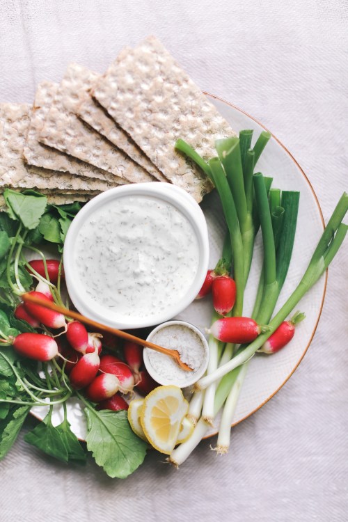 A plate with crispbread, fresh radishes, green onions, lemon, and a bowl of creamy dip garnished with herbs.
