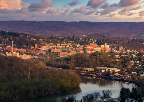 A scenic view of a town by a river, surrounded by hills and trees, under a colorful sky at sunset.
