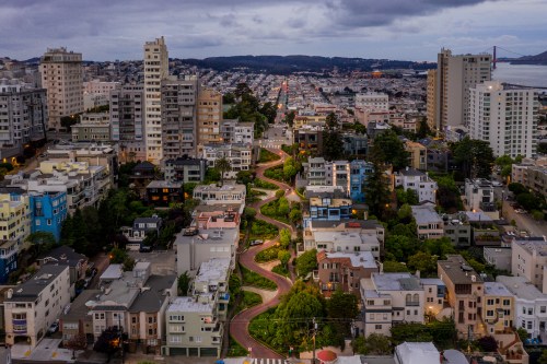 Aerial view of a winding street surrounded by buildings and greenery in a cityscape, with distant hills and cloudy sky.