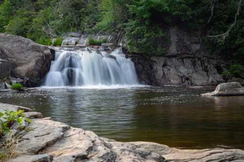 A serene waterfall cascades into a calm pool, surrounded by lush greenery and rocky terrain.