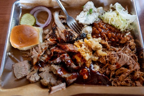 A tray filled with various barbecue meats, sides of baked beans, coleslaw, pickles, onions, and a bun.