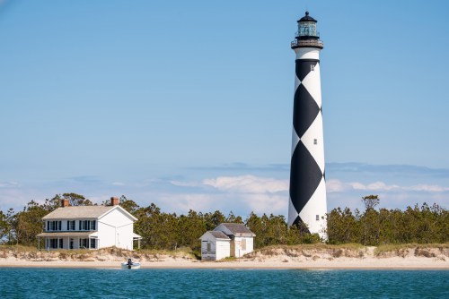 A black and white striped lighthouse stands beside a white house on a sandy shore, with calm blue waters in the foreground.