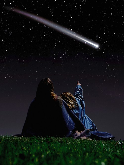 Two people wrapped in a blanket gaze at a comet streaking across a starry night sky.