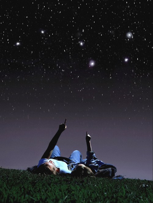 Two children lie on a grassy field, pointing at stars in a clear night sky filled with twinkling stars.