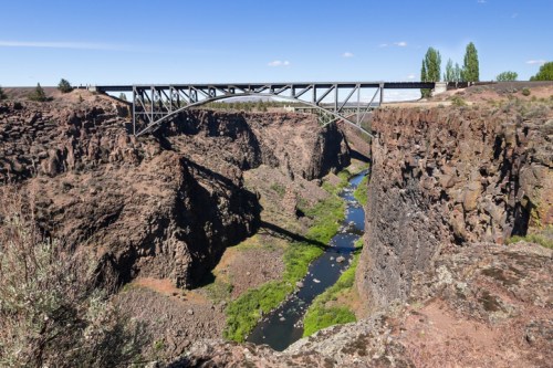 A metal bridge spans a deep canyon with a winding river below, surrounded by rocky cliffs and sparse vegetation.