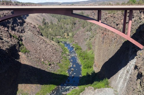 A modern bridge spans a deep canyon, casting a shadow over a winding river surrounded by rocky cliffs and greenery.