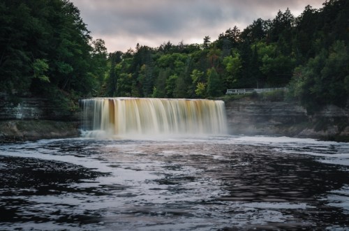 A serene waterfall cascades into a river, surrounded by lush green trees under a cloudy sky.