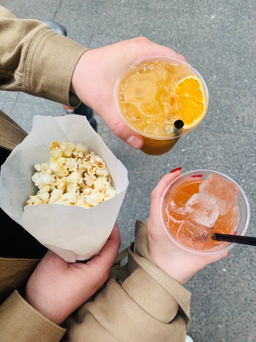 Two hands hold drinks and a bag of popcorn, with one drink garnished with an orange slice.