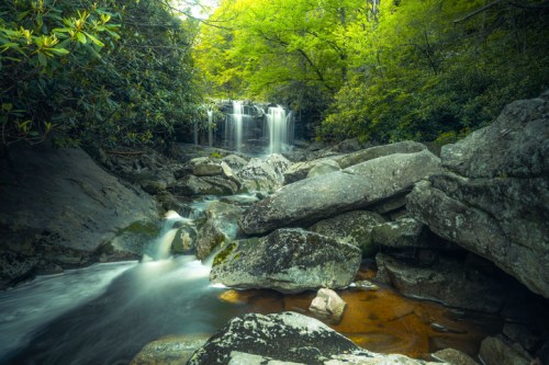 A serene waterfall cascades over rocks, surrounded by lush green trees and a tranquil stream.