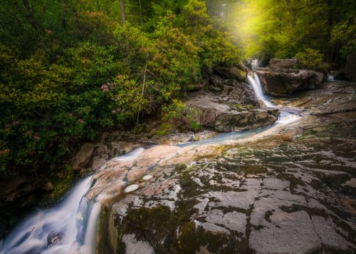 A serene waterfall cascades over rocks, surrounded by lush greenery and soft sunlight filtering through the trees.