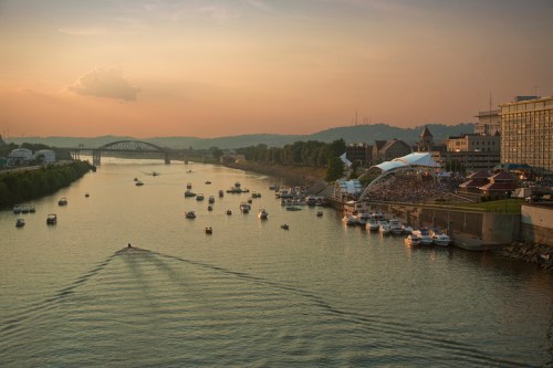 A sunset view over a river filled with boats, with a crowd gathered along the shore and a bridge in the background.
