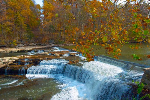 A serene waterfall cascades over rocks, surrounded by autumn foliage in vibrant orange and yellow hues.