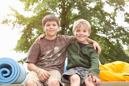 Two boys sit together outdoors, smiling, with face paint and casual clothing, surrounded by colorful mats and a tree.