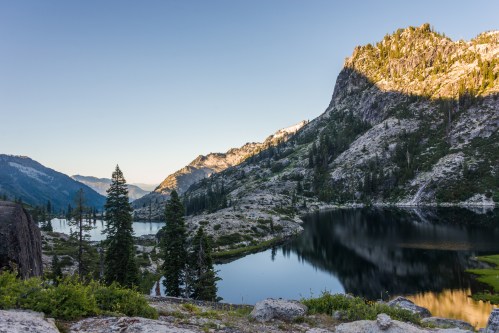A serene mountain landscape with a calm lake, surrounded by trees and rocky terrain under a clear blue sky.
