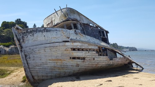 An old, weathered boat rests on a sandy shore, partially submerged, with trees and houses in the background.