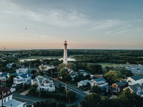 Aerial view of a coastal town with a lighthouse, surrounded by trees and houses, under a clear sky at sunset.