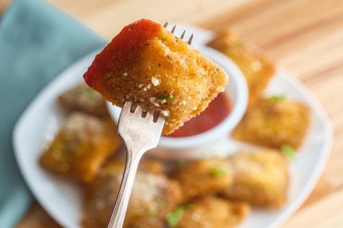 A fork holding a crispy, golden-brown ravioli topped with marinara sauce, with more ravioli in the background.