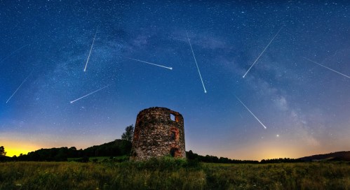 A starry night sky with shooting stars over a stone tower in a grassy field, surrounded by trees and distant hills.
