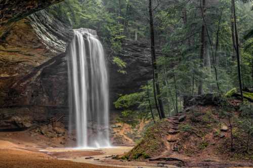 A serene waterfall cascades down rocky cliffs, surrounded by lush green trees in a tranquil forest setting.