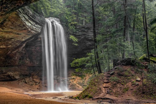 A serene waterfall cascades down rocky cliffs, surrounded by lush green trees in a tranquil forest setting.