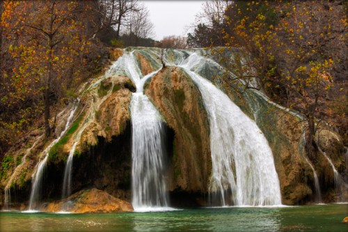 A serene waterfall cascading over rocky cliffs, surrounded by autumn foliage and a calm green pool below.