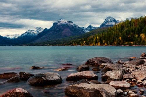 A serene lake surrounded by mountains, with rocky shores and autumn trees under a cloudy sky.