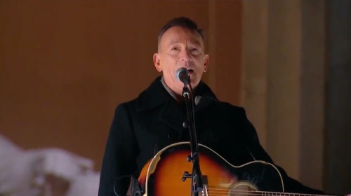 A musician performs on stage with a guitar, speaking into a microphone, wearing a black coat.